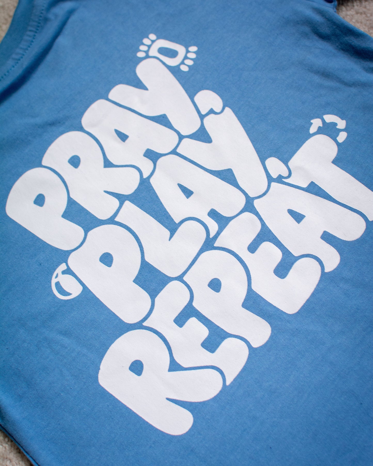 Blue t-shirt with white text 'PRAY, PLAY, REPEAT' on a gray surface